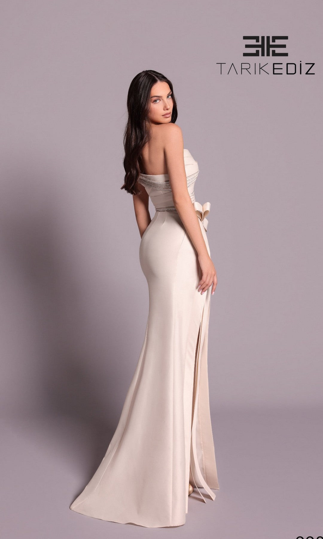 A woman models the Tarik Ediz 99093 long formal dress, a strapless cream gown, posing with her back slightly turned. The plain background displays the